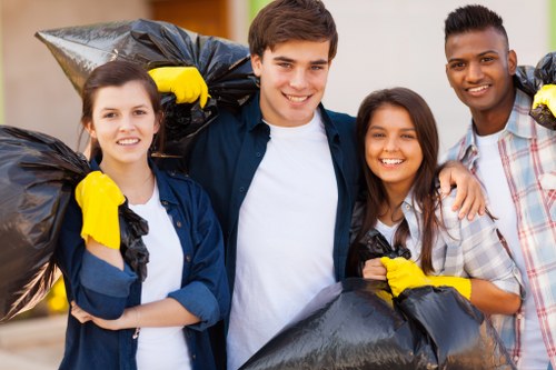 Four young volunteers outdoors, each holding large black rubbish bags filled with collected waste. The group includes two women and two men, all smiling and wearing casual clothing suitable for outdoor activity. Some are wearing yellow gloves, indicating active participation in rubbish collection or community clean-up. They stand on a paved surface with a blurred background of greenery and buildings, suggesting a local neighbourhood or community space. The black rubbish bags are visibly full and tied at the top, with their materials and textures indicating plastic. Their expressions and posture convey a sense of teamwork and community engagement, aligned with the typical context of rubbish removal efforts in Barking or surrounding areas. This scene captures the essence of community clean-up activities, which local rubbish removal services like Office Clearance Barking often support, demonstrating a shared commitment to maintaining clean environments.