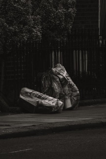 Two large black trash bags filled with waste are placed on the pavement beside a low curb, leaning slightly against a black metal fence with vertical bars. The bags appear to be made of thick plastic 